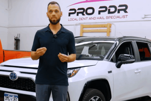 A man in a navy polo shirt stands in front of a white Toyota SUV, speaking to the camera inside a repair shop with a “PRO PDR Local Dent and Hail Specialist” sign on the wall behind him.