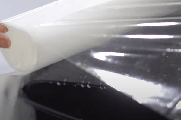 clear paint protection film (PPF) being carefully applied to a glossy black car surface, showing the film's flexibility and smooth adhesion as it shields the paint beneath.