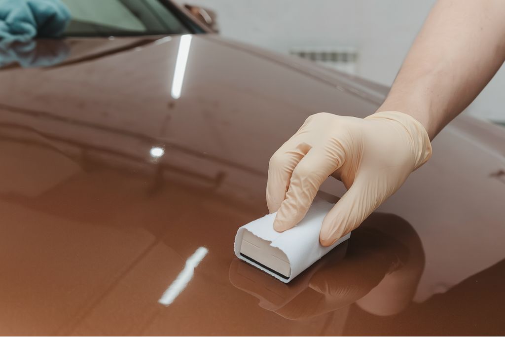 A gloved hand applies ceramic coating to a brown car hood using an applicator pad, showing the beginning stage of how ceramic coating works by bonding to the paint for long-term protection.