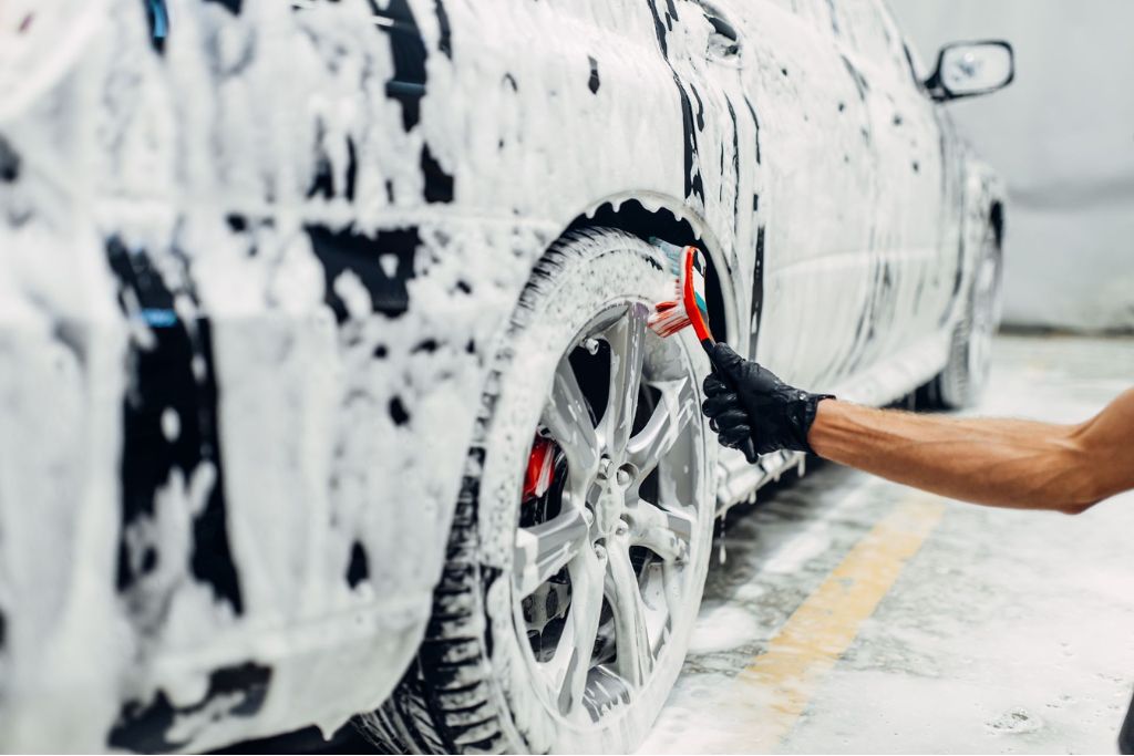 A gloved technician cleans a car’s wheels with a detailing brush during the initial wash phase, an essential part of the ceramic coating application process to ensure proper bonding and a flawless finish.