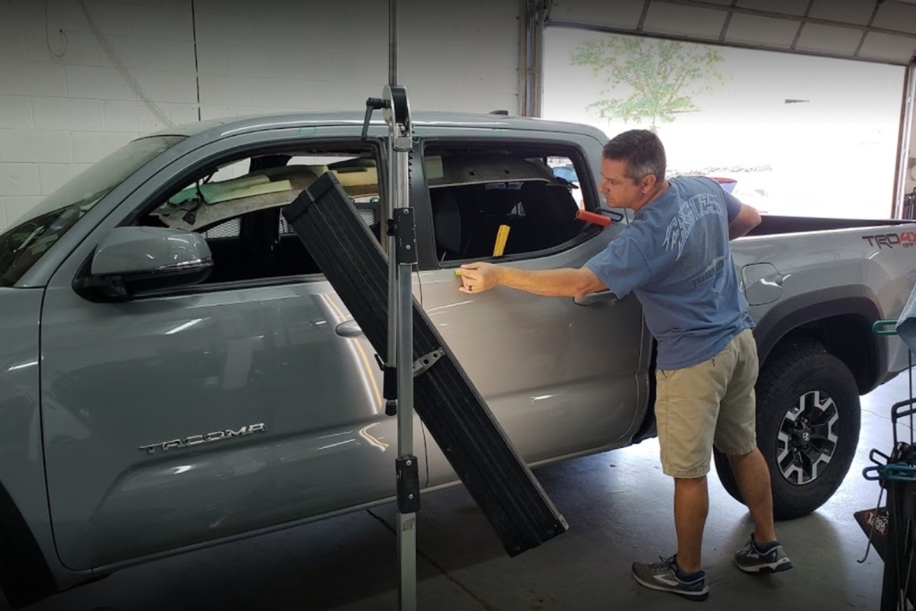 Technician performing paintless dent repair on a silver Toyota Tacoma using a reflection board and hand tools to access a dent on the rear door, demonstrating how paintless dent repair works and when it may or may not be effective.