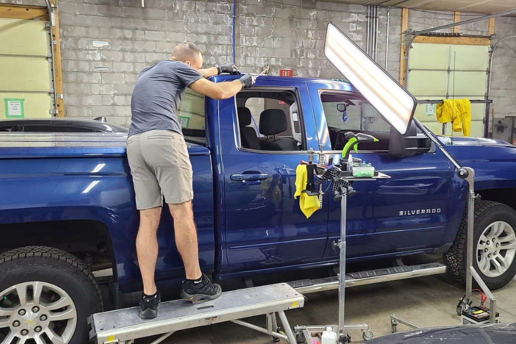 Technician standing on a platform while performing paintless dent repair on a blue Silverado, using reflection lights and specialized tools to access and reshape the dented panel from behind during the paintless dent repair process.