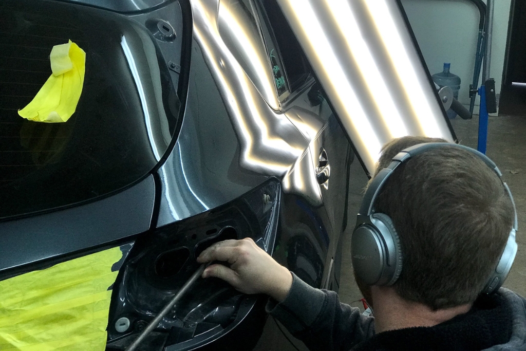 Hail Damage Repair Process shown as a technician in Colorado Springs performs paintless dent repair on a dark-colored vehicle using specialized tools and reflection lighting. The image highlights the precision and non-invasive nature of PDR to restore hail-dented panels.