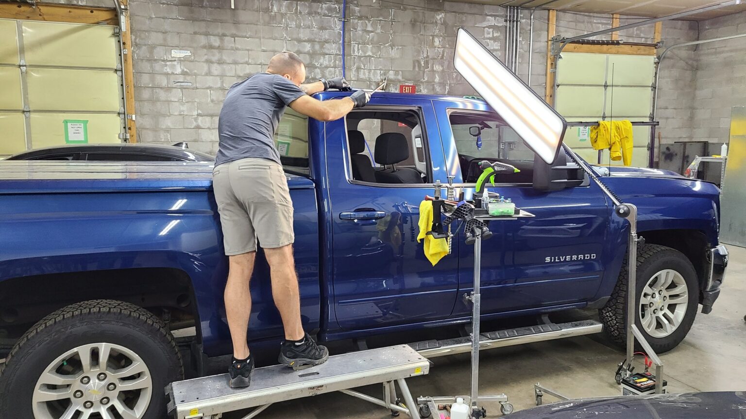 Technician performing paintless dent repair on a blue Chevrolet Silverado truck in an auto repair shop, with tools and equipment visible for hail damage repair.