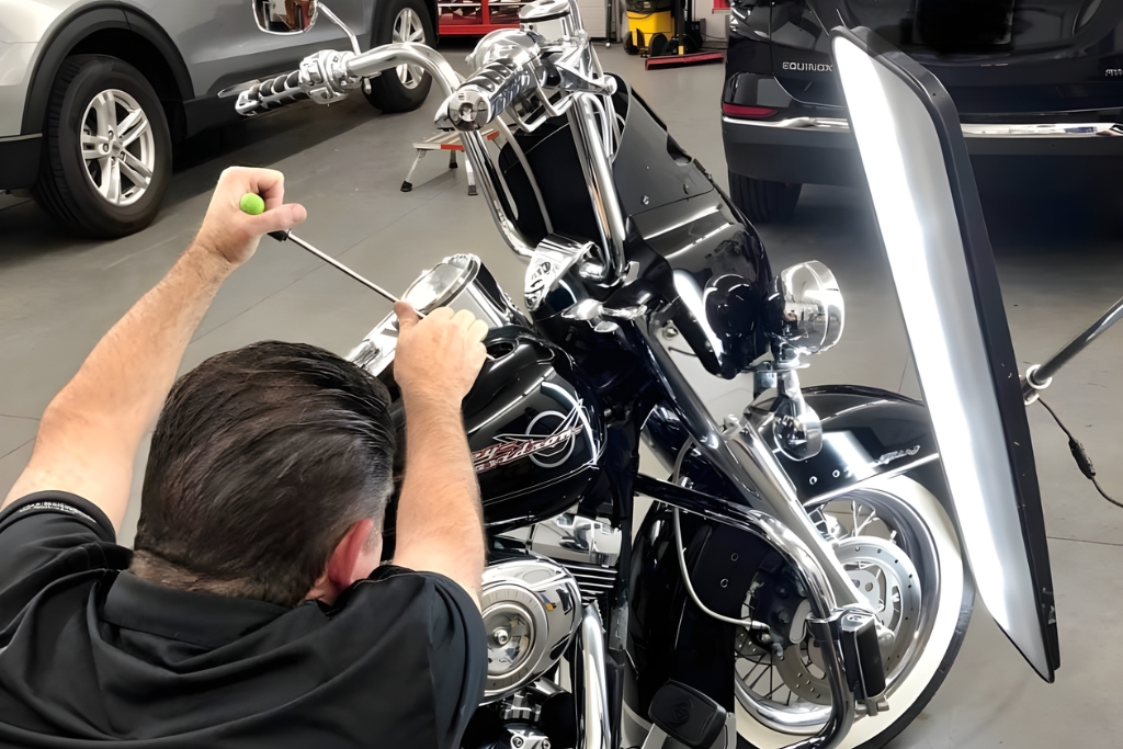 A technician uses a precision rod and reflection board to perform paintless dent repair on a black Harley-Davidson motorcycle in a professional shop, illustrating the detailed motorcycle PDR process used in Colorado Springs.