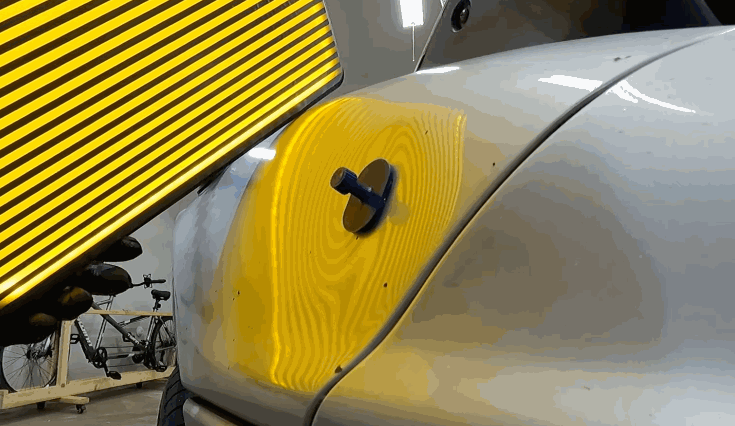 Technician using a yellow-striped reflection board to align a glue pull tab on a car panel during a paintless dent repair process.