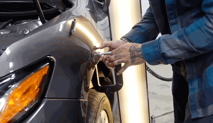 A technician using a metal tool to precisely press out a dent from a dark-colored vehicle’s fender during paintless dent repair.