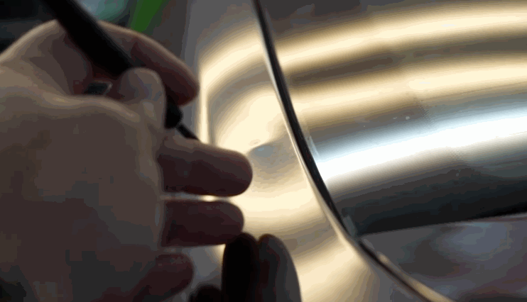 Technician marking a small dent on a car's surface under reflective lighting during hail damage repair inspection.
