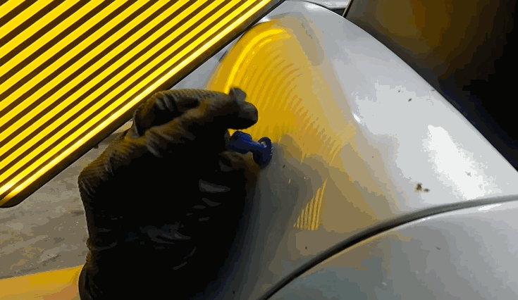 Technician wearing black gloves applying a glue tab to a dented car panel during the paintless dent repair process, with a yellow-striped reflection board highlighting the dent.