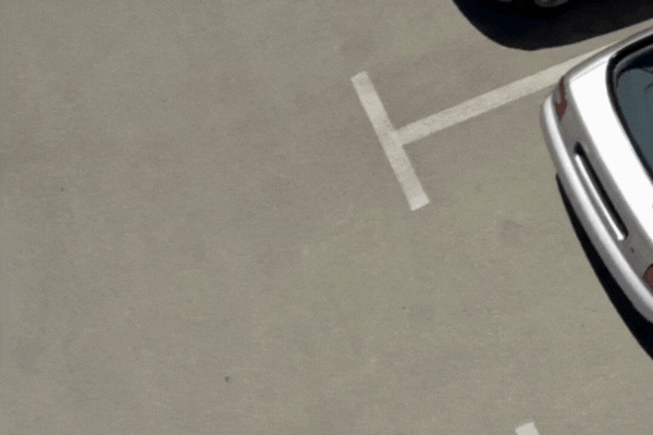 White sedan reversing into a clearly marked parking spot, grazing the bumper of another car in a low-speed collision typical of a fender bender.