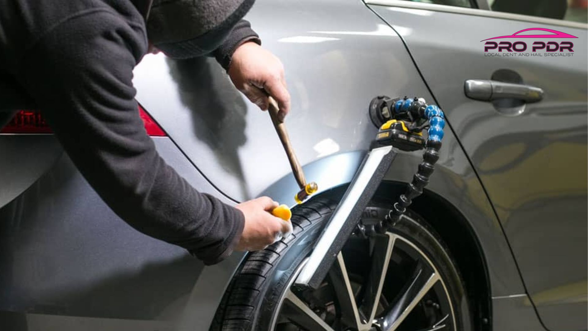 Technician taps out a dent with a hammer and plastic tool while a light reflector and suction device assist in paintless dent repair on a silver car’s rear fender.