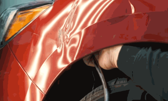 Technician performing fender bender repair by using a rod to push out a dent from the inside of a red car’s fender.