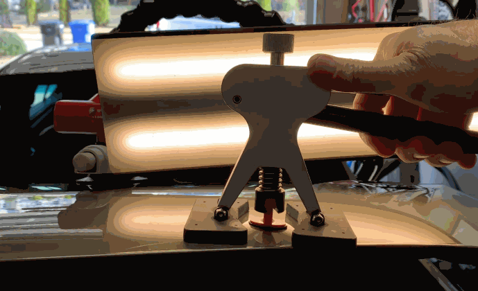 Paintless dent repair (PDR) tool being used on a car hood dent under LED reflection lighting to highlight imperfections.