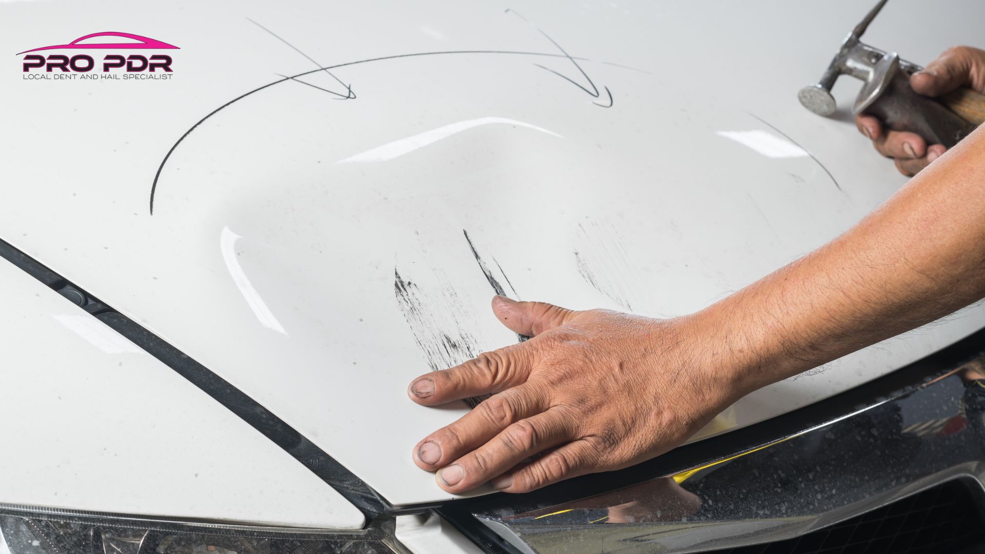 Close-up of a person inspecting and beginning to repair a large car hood dent with visible scratches on a white vehicle.