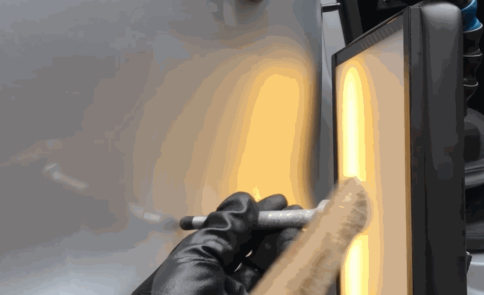Gloved hand adjusting a dent removal tool during Paintless Dent Repair as a reflection light panel reveals surface distortions on a vehicle door.