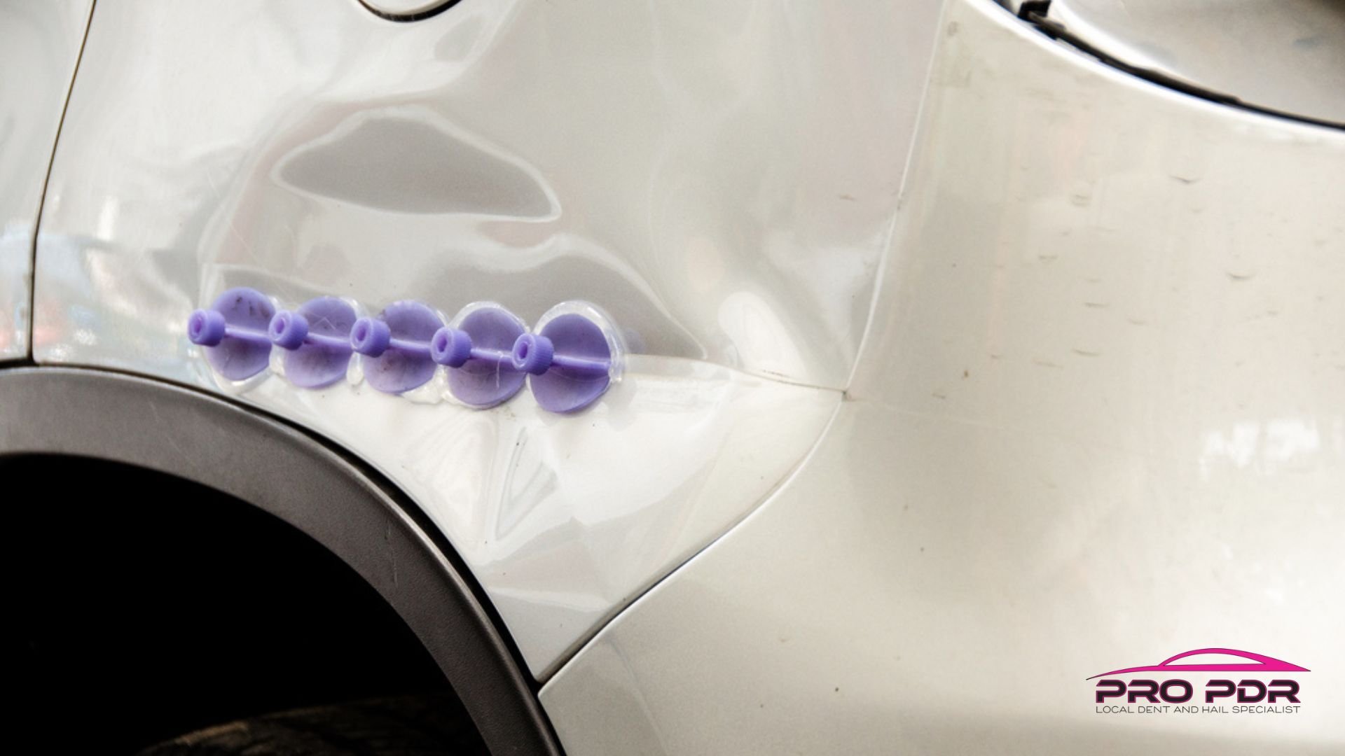 White car panel with a visible dent being repaired using a row of purple suction tools as part of paintless dent repair. The setup shows PDR in progress on the vehicle exterior without affecting the paint.