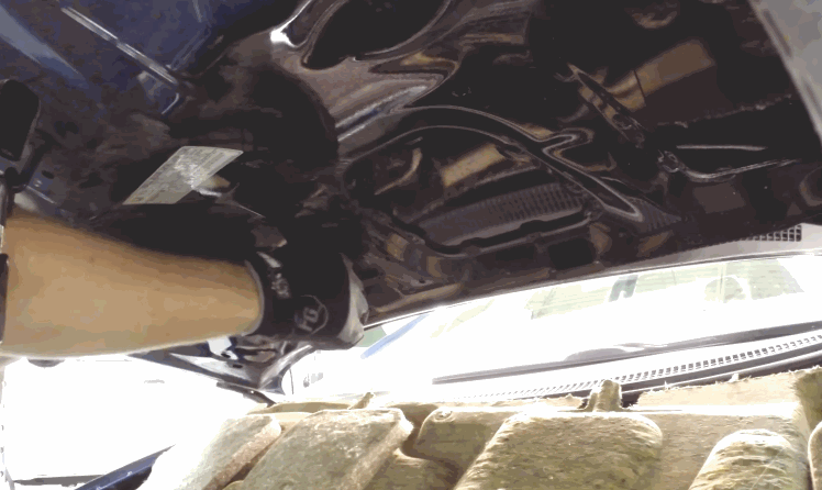 A technician wearing gloves works underneath a car hood, accessing the underside of the panel to repair hail damage dents from within. The interior metal structure is visible as tools are positioned for precision work.