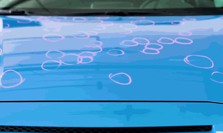 Blue car hood covered in circular reflections and highlighted marks showing areas of hail damage across the surface. The pattern of dents is emphasized by overhead lighting to reveal uneven metal.