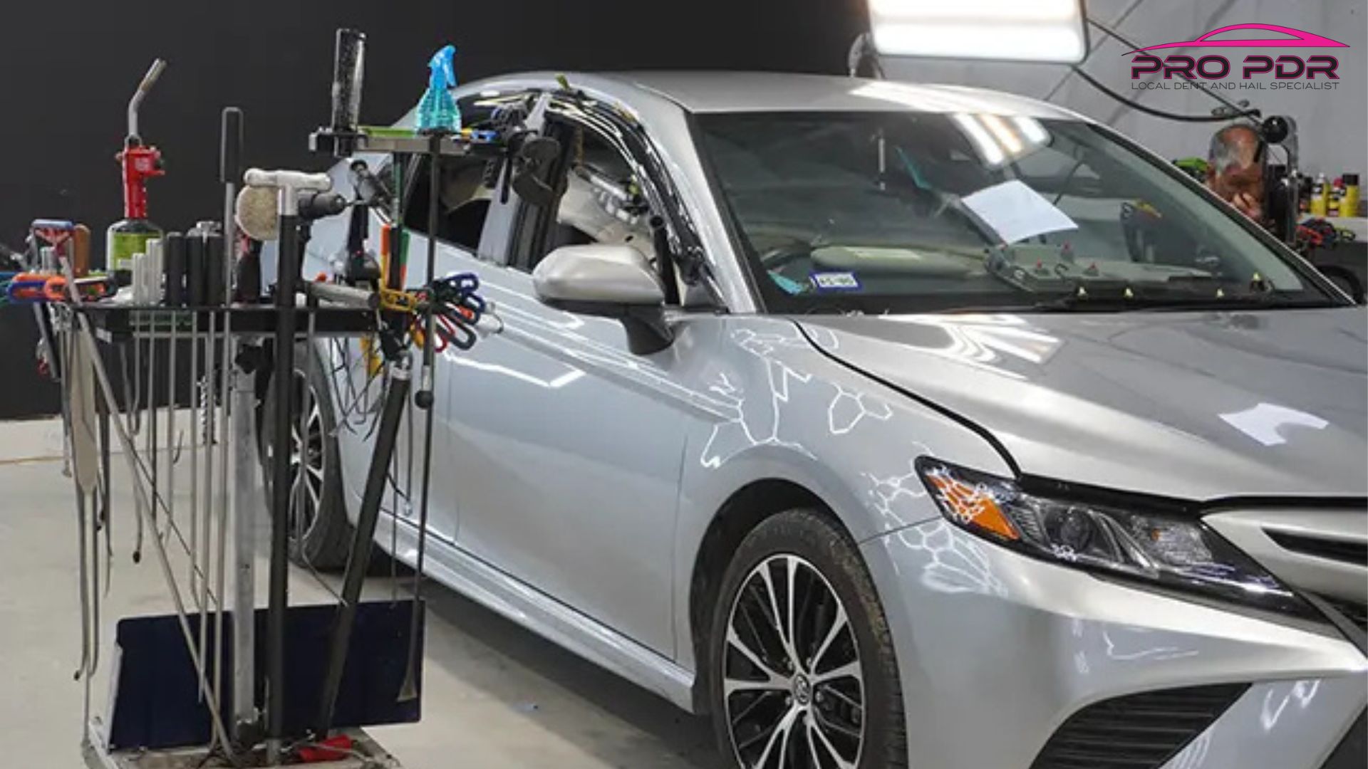 A silver car sits inside a repair shop beside paintless dent repair tools for hail damage repair. The upper corner includes the text "PRO PDR LOCAL DENT AND HAIL SPECIALIST".