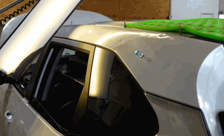 A silver vehicle roof with multiple dents is set up for hail damage repair under bright shop lighting. Green pads and small tabs are placed on the panel to prepare the damaged area for dent removal.