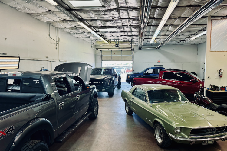 Automotive service garage interior featuring various vehicles including a classic green Mustang, a gray truck, and a black Ford SUV, highlighting Pro PDR's extensive vehicle repair and surface protection capabilities.