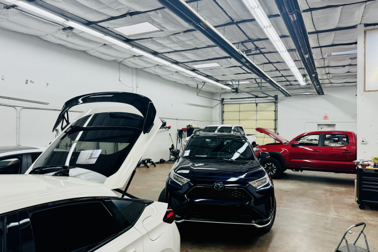Automotive repair shop interior featuring multiple vehicles, including a white car with an open trunk, a black SUV, and a red truck, showcasing a clean and organized workspace for paintless dent repair and surface protection services.